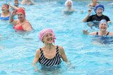 An elderly woman in a red and white swim cap smiles in a swimming pool with other women behind her.