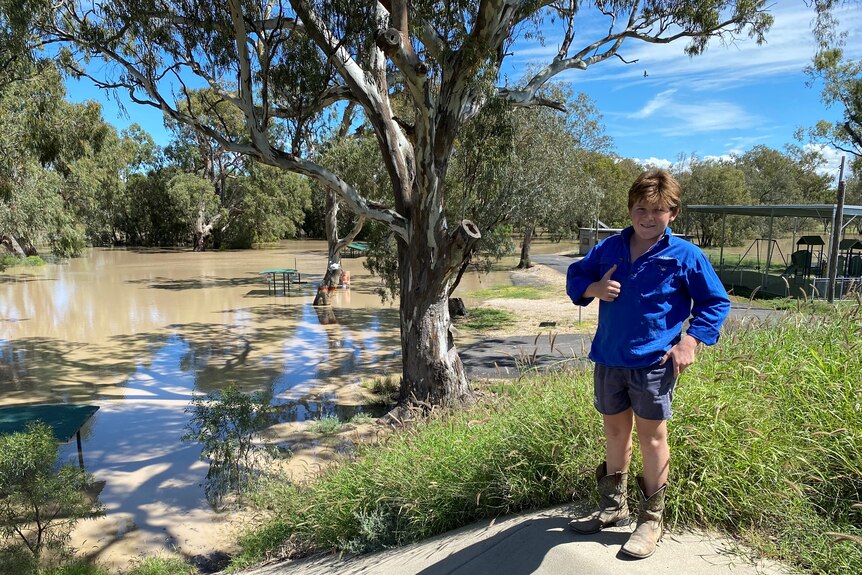 Boy in blue shirt smiles and gives a thumbs up while standing next to a full river.