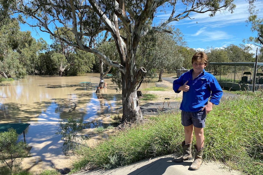 Boy in blue shirt smiles and gives a thumbs up while standing next to a full river.