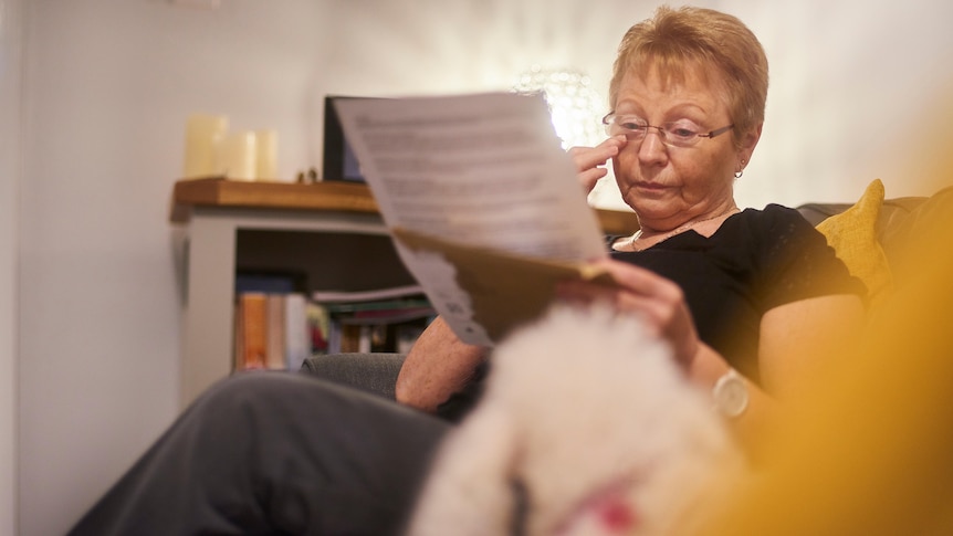 older woman wearing glasses in a chair reading a letter who appears upset