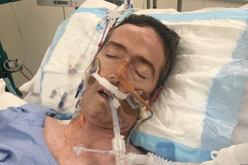 Man lies in hospital bed with tubes attached to his chest and face