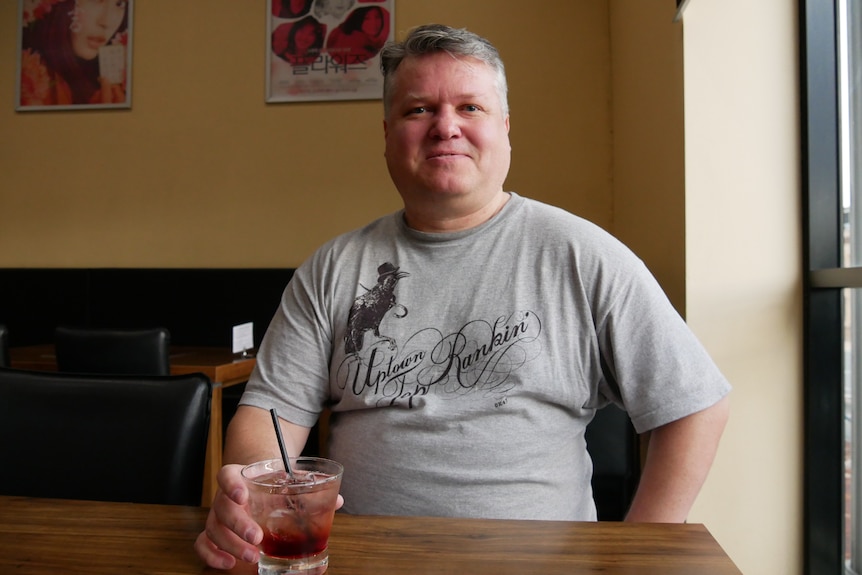 a man in a grey shirt sits at a restaurant table with a drink 