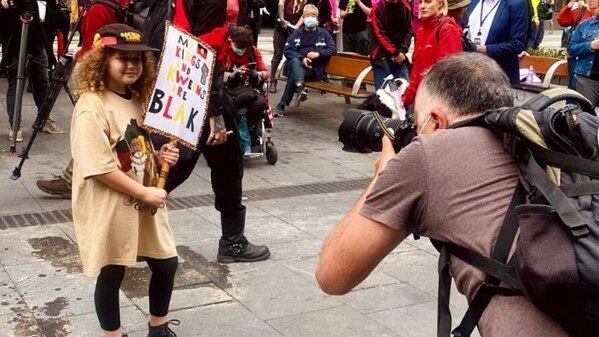 Young girl holding a sign that reads "My Kings and Kweens are Blak", with a man holding a camera taking a photo of her.