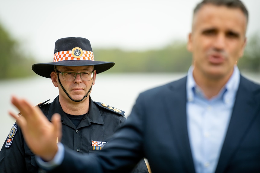 The SES CE in uniform with a hat standing behind the premier of South Australia in a press conference 