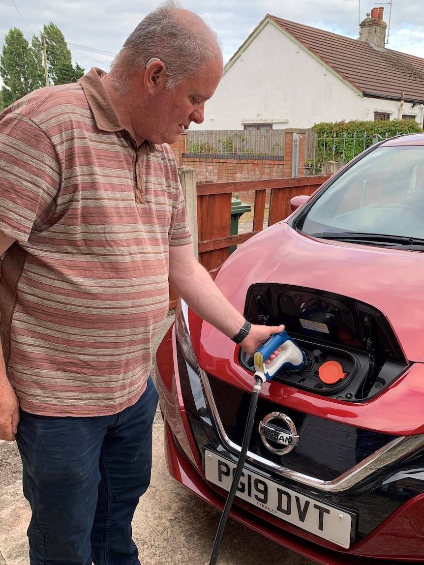 An elderly man plugs a charging cord into the front of an EV