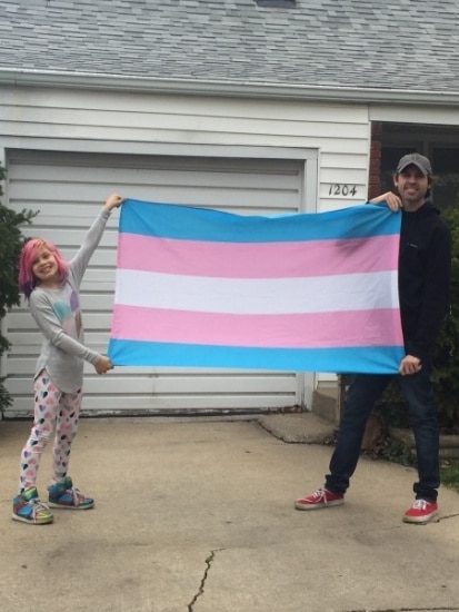 A young girl an a man stand in front of a white house, holding a blue, pink and white striped flag