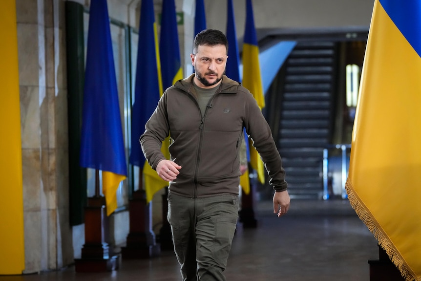 Zelenskyy walks in front of blue and yellow Ukrainian flags indoors.