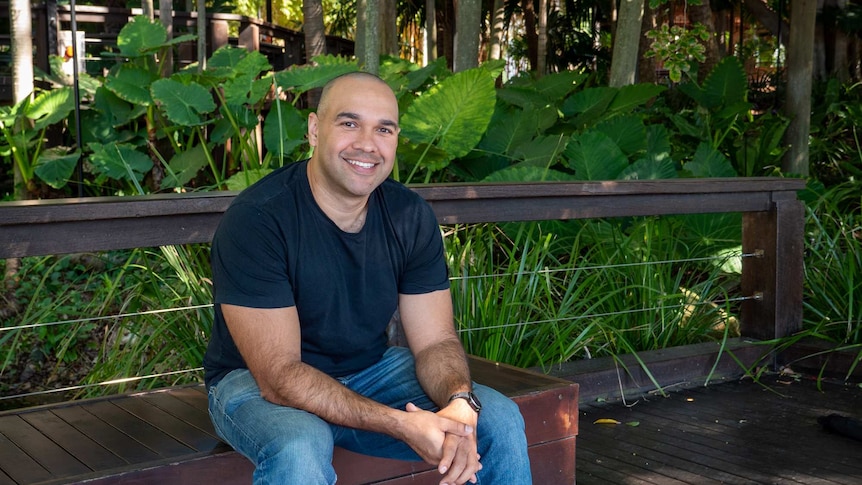 Craig Quartermaine sitting on a wooden bench with tropical plants in in a lush garden behind him.