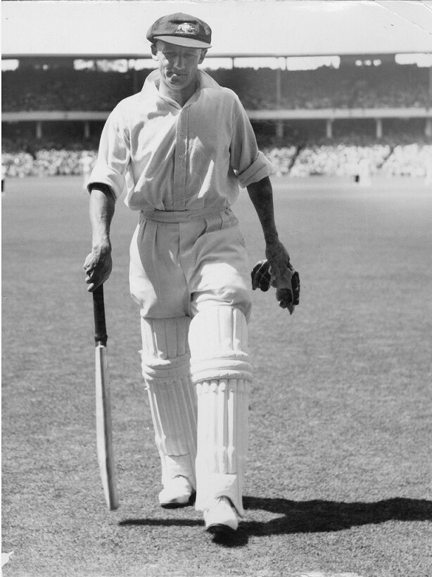 Don Bradman walking off with bat and gloves in his hands, baggy green on his head.