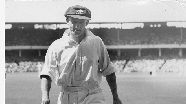 Don Bradman walking off with bat and gloves in his hands, baggy green on his head.