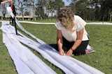 A woman kneeling on the ground sewing lengths of white knitting together.