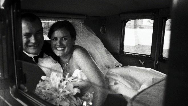 A black and white photograph of a bride and a groom in a car at their wedding day.