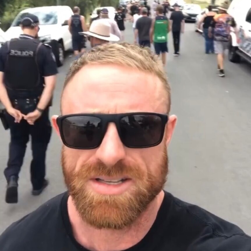 A close up of a man's face wearing sunglasses with a crowd of people and police walking behind him.