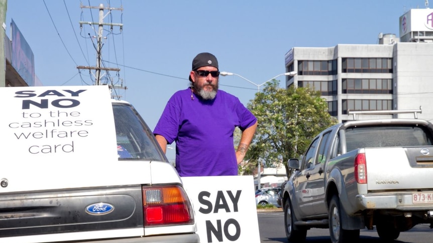 A man stands beside his car with a sign reading "say no to chashless welfare".