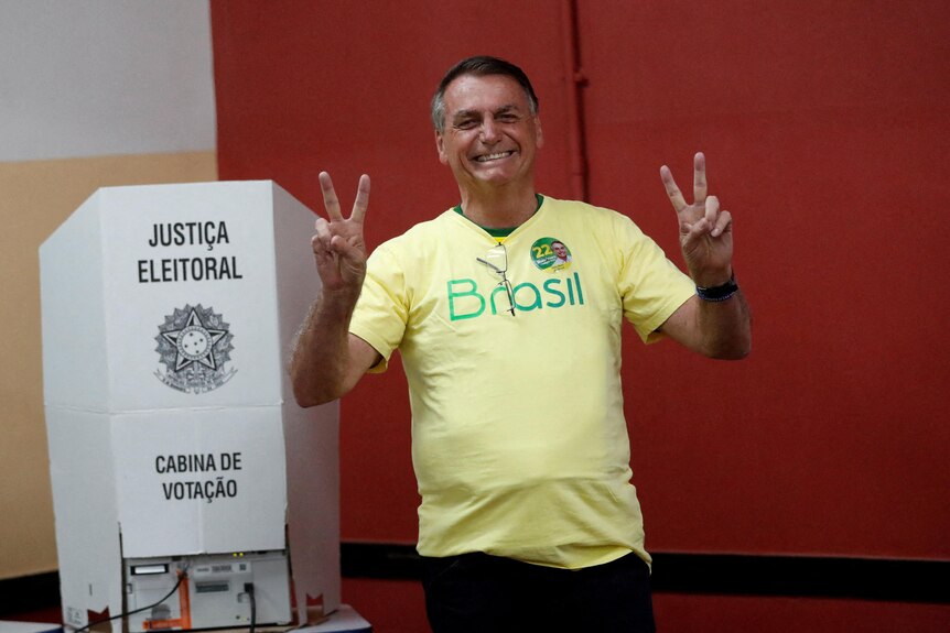Jair Bolsonaro dos peace signs with his hands standing in front of a white cardboard voting booth.