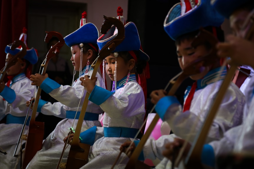 A row of children with blue hats play stringed instruments, with looks of concentration on their faces