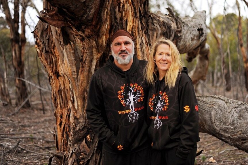 A man with a grey beard and a bandana stands with a blonde-haired woman in front of a huge tree