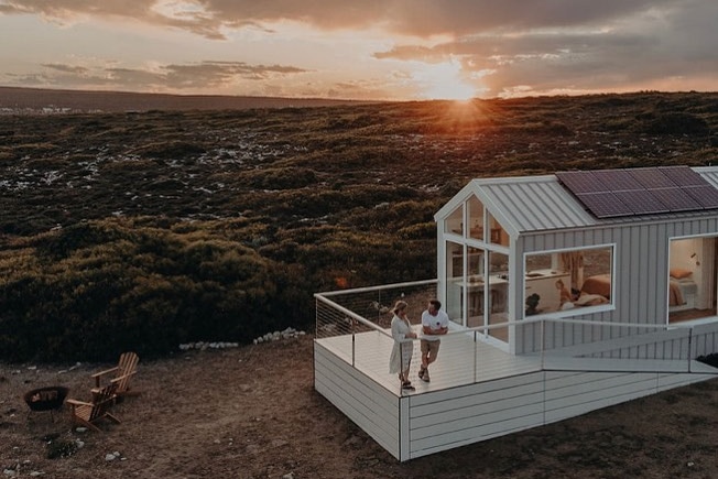 Two people stand on the front deck of a tiny home with a sunset backdrop.