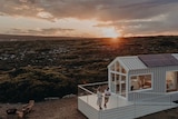 Two people stand on the front deck of a tiny home with a sunset backdrop.