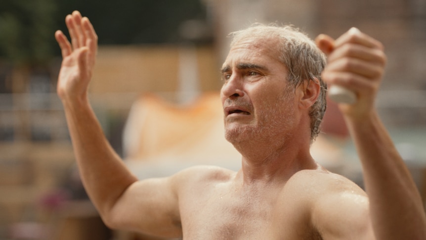 A film still of a shirtless Joaquin Phoenix looking distressed. He stands with his arms up, one hand clutching an ornament.