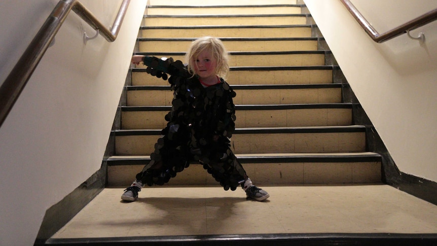 A young girl stands on the landing of a staircase dressed in a black, sparkling onesie.