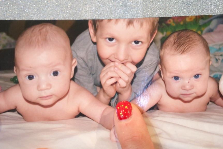 a young boy smiles at the camera, sitting in the middle of two baby twins