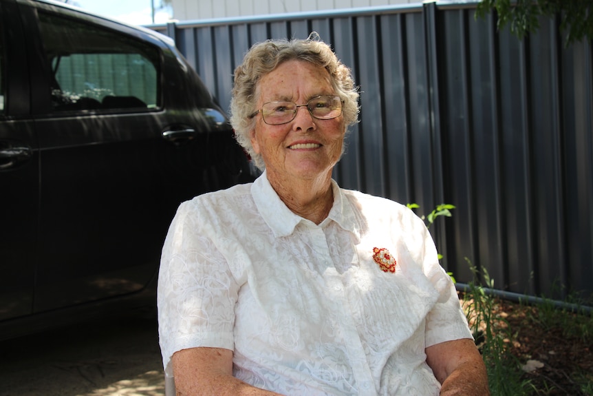An older woman in a white shirt with a red brooch smiles, a dark car and fence behind her