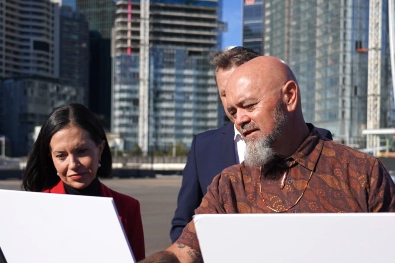 A woman and two men stand in front of plans on stands with buildings in the background