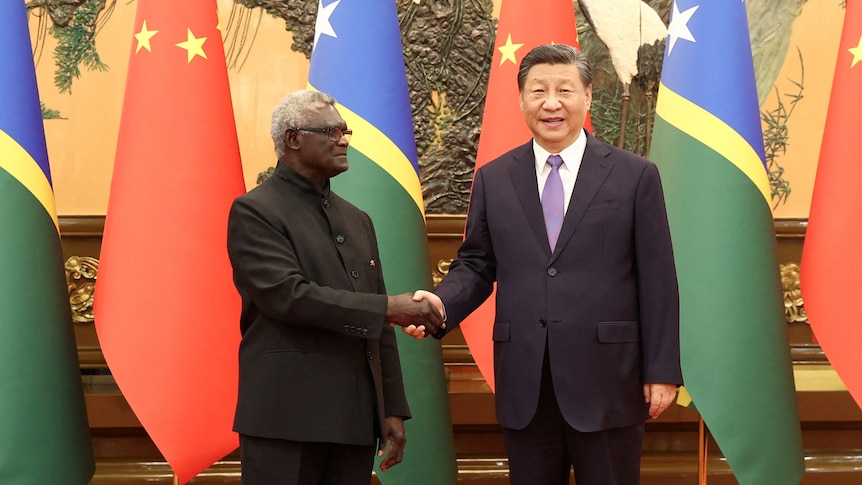 Two men shake hands with red china and blue green Solomon Islands flags in the background.