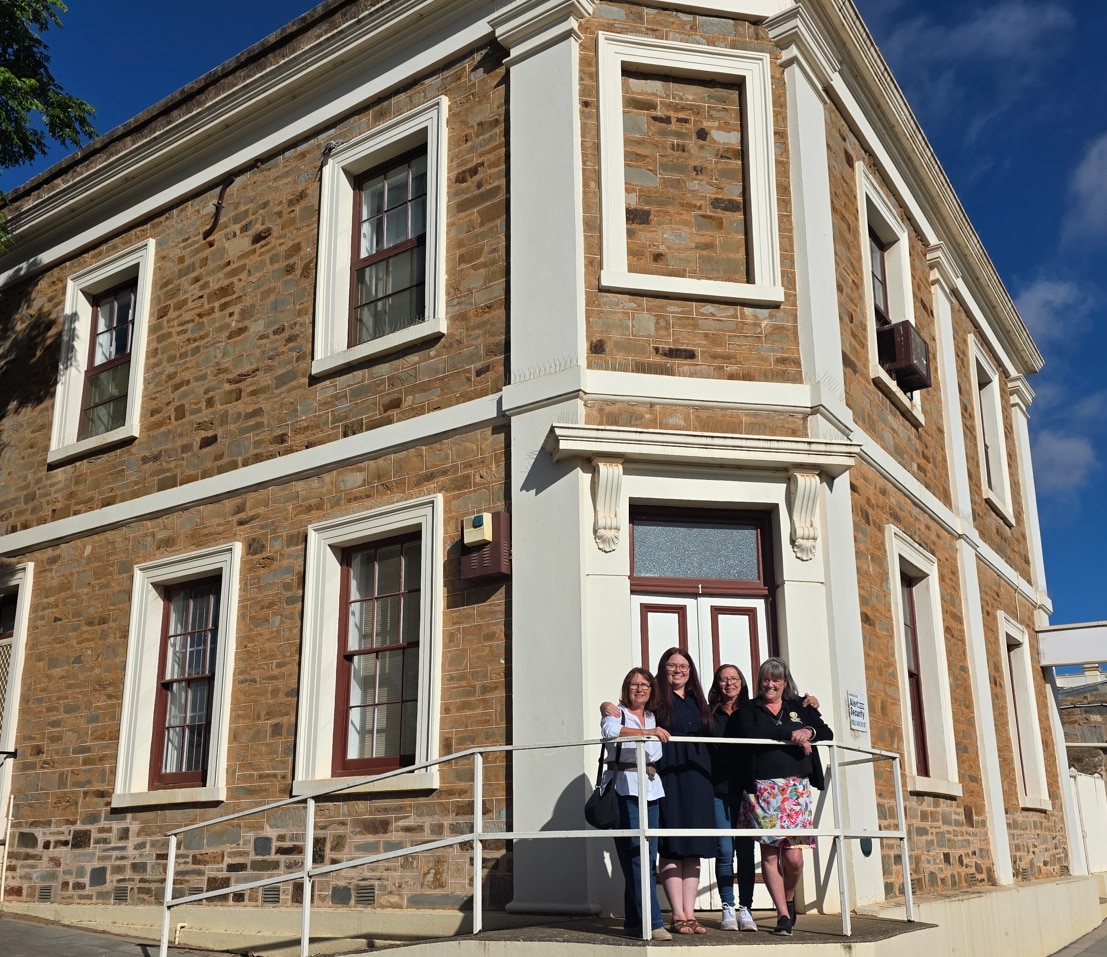 A two story sandstone building on a street corner with four people standing in front of it