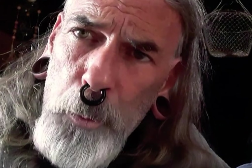 A man with stretchers, a septum ring and long grey hair and a beard.