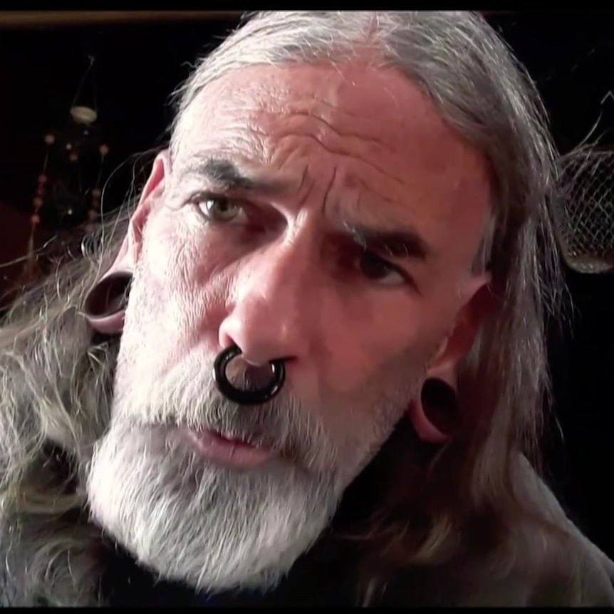 A man with stretchers, a septum ring and long grey hair and a beard.