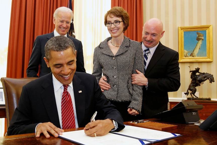 Barack Obama signing a piece of paper, while Gabby Giffords, Mark Kelly and Joe Biden look on smiling