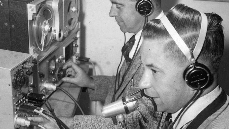 A man wearing headphones speaks into a microphone with a wire guard attached. Tape operator in background.