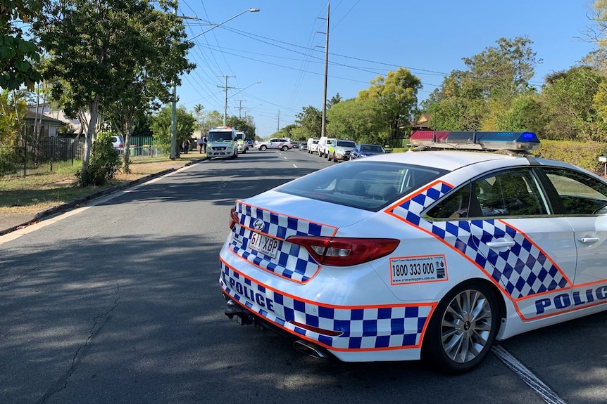 A police car blocking the road and a number of ambulances and police cars parked along the road in a suburban street