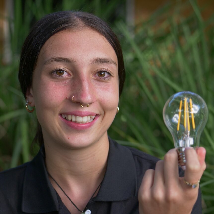 Young girl with brown hair slicked back into a bun smiles while holding a lightbulb.