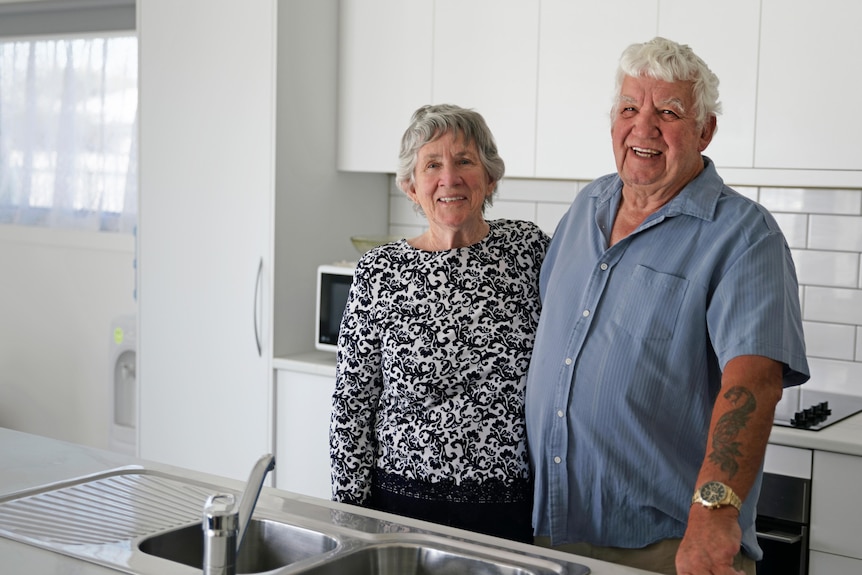 Maria and Peter stand together in their kitchen smiling. They have grey hair.