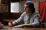 A white man in his 30s with brown hair, wears a backwards baseball cap and a grey checkered shirt and sits at a table speaking.