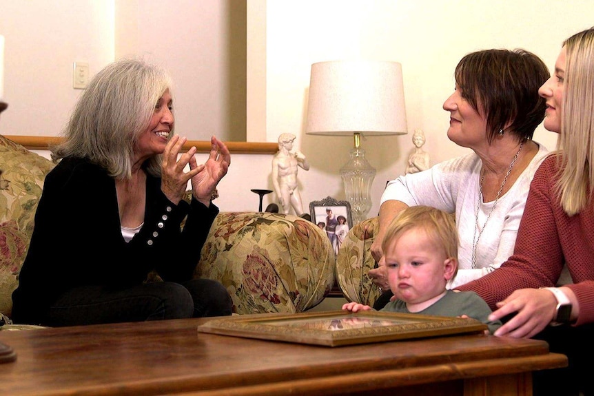 Three women sit together talking. A baby is looking at a photo in front of them.