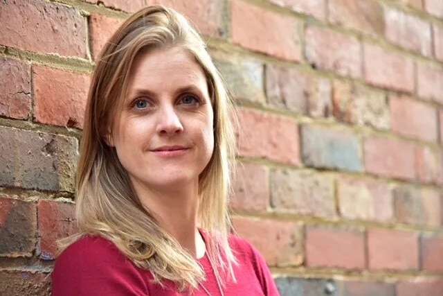 A portrait of a woman with blonde hair leaning against a wall 