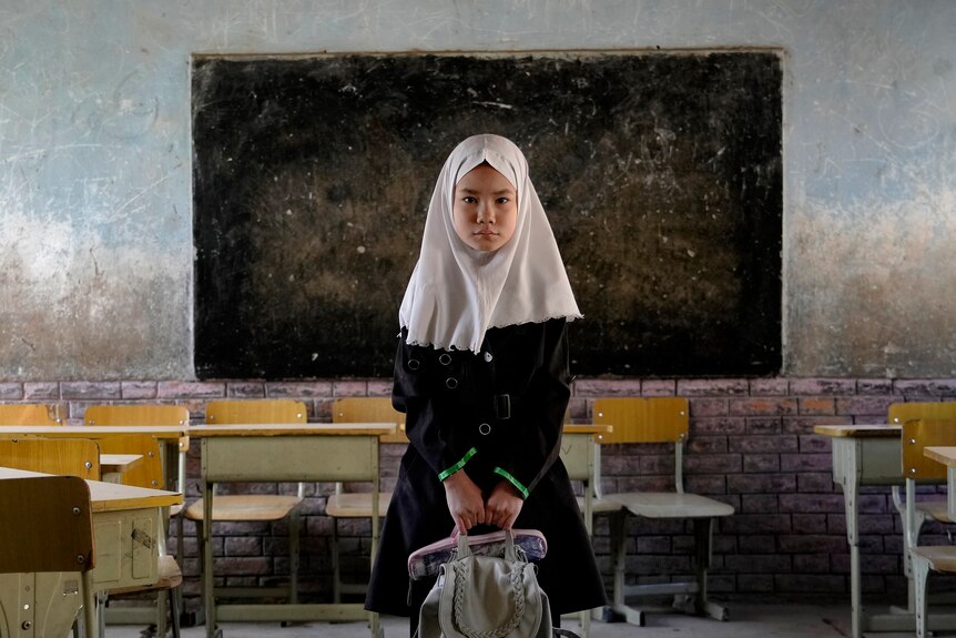 A young girl wearing a headscarf and holding a school bag poses for a photo in her classroom.
