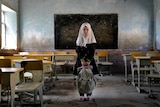 A young girl wearing a headscarf and holding a school bag poses for a photo in her classroom.