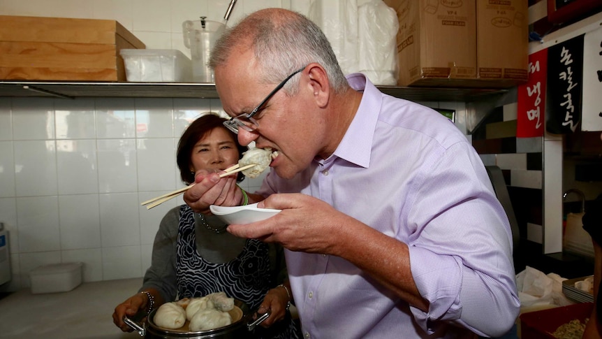 Scott Morrison eats a dumpling with a pair of chopsticks. His face is scrunched up and the bowl is close to his face.
