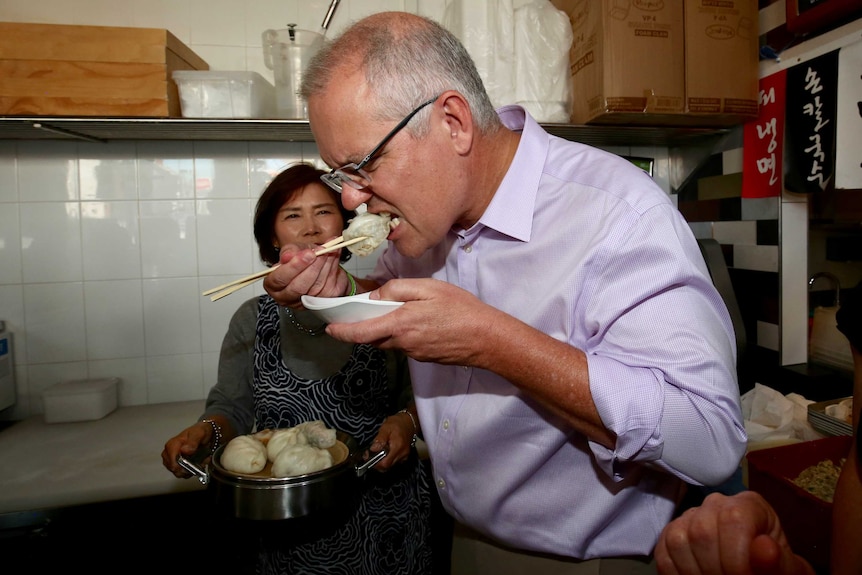 Scott Morrison eats a dumpling with a pair of chopsticks. His face is scrunched up and the bowl is close to his face.