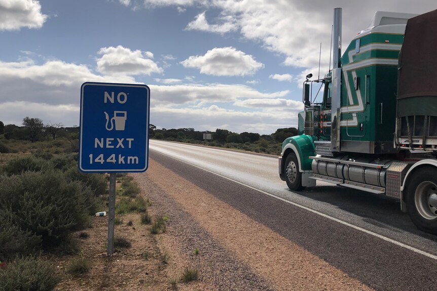 Sign saying No Fuel For 144km, at road's edge green semi-trailer truck passing on the right 