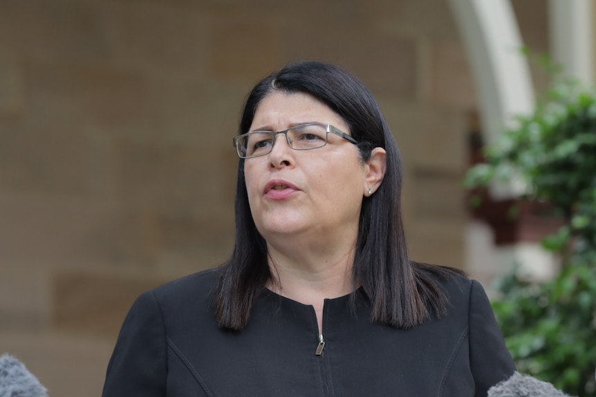 A w0oman with dark should-length hair and thin framed glasses speaks at a lecturn while wearing a black dress. 