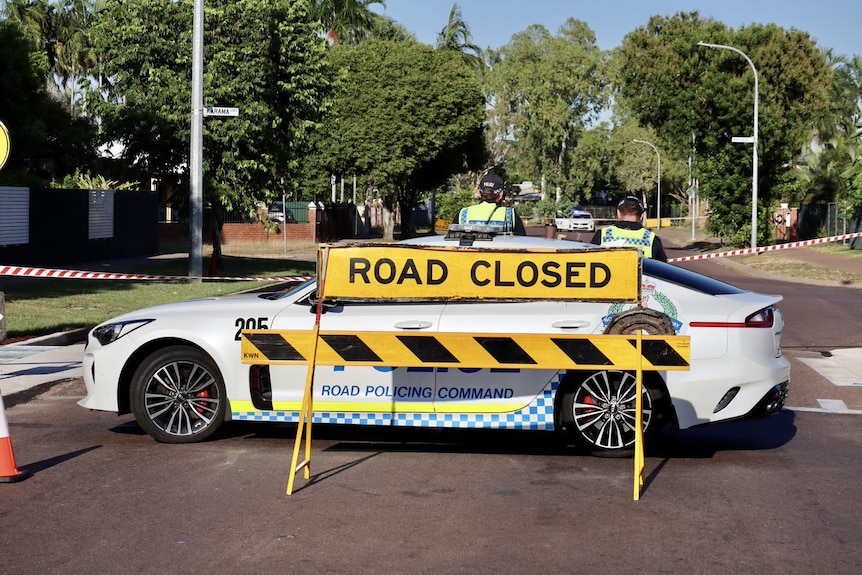 A neon yellow sign reading road closed stands in front of a police car on a street in Darwin. 
