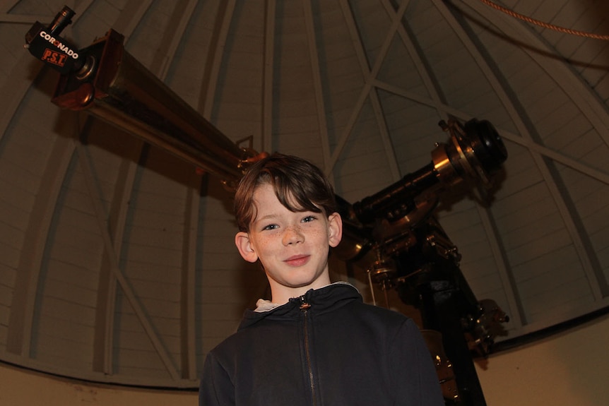 A smiling 11-year-old boy stands in front of a large telescope inside a domed roof.