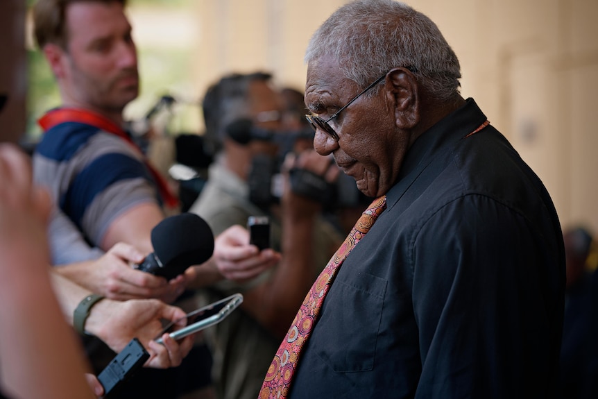 A man in a black shirt and pink tie talks into microphones.