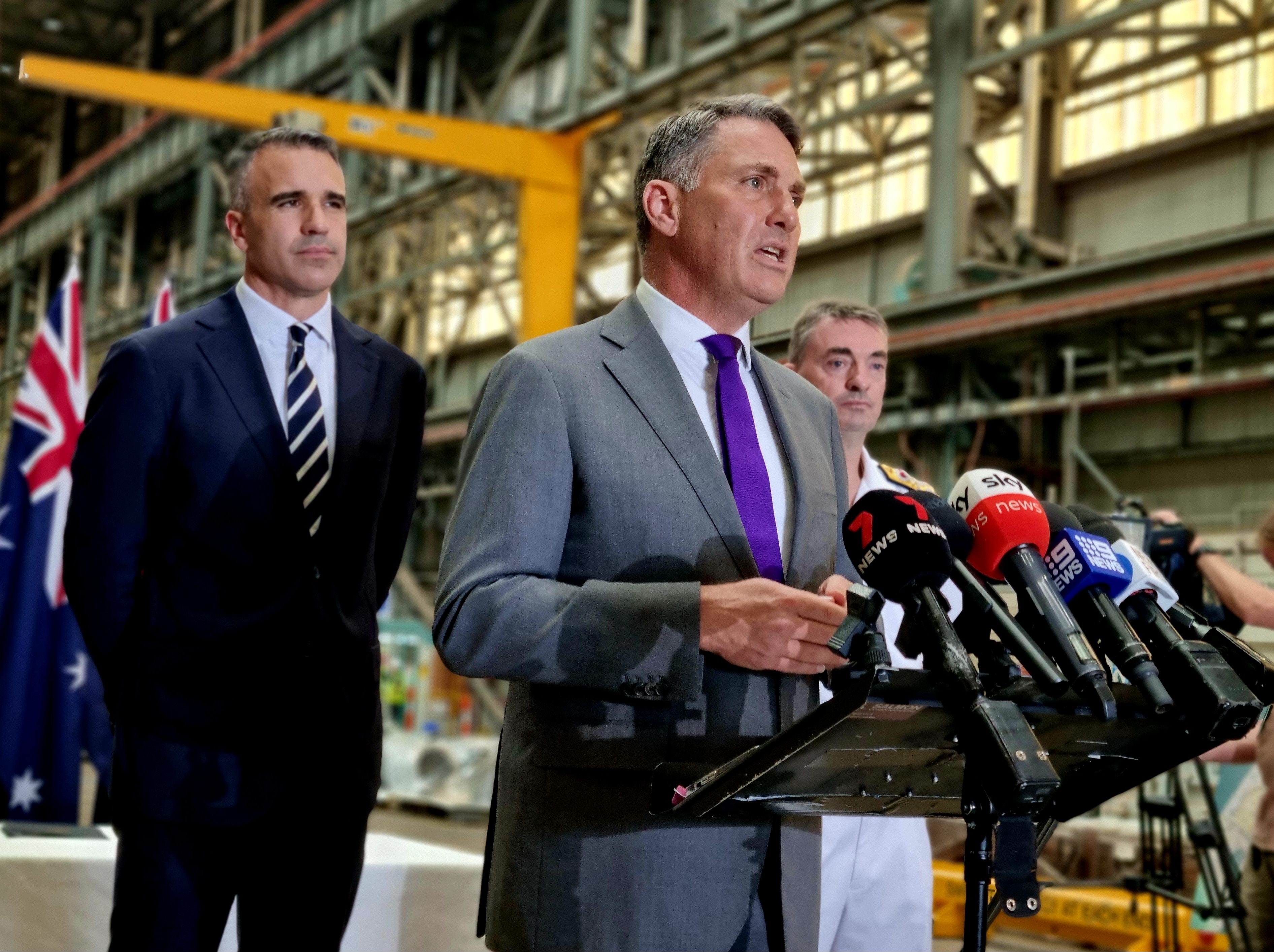 One man speaks in front of media microphones, with two men standing behind him on either side